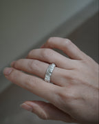 A close-up of a hand wearing a textured silver band ring with small clear stones, set against a soft, out-of-focus neutral background.