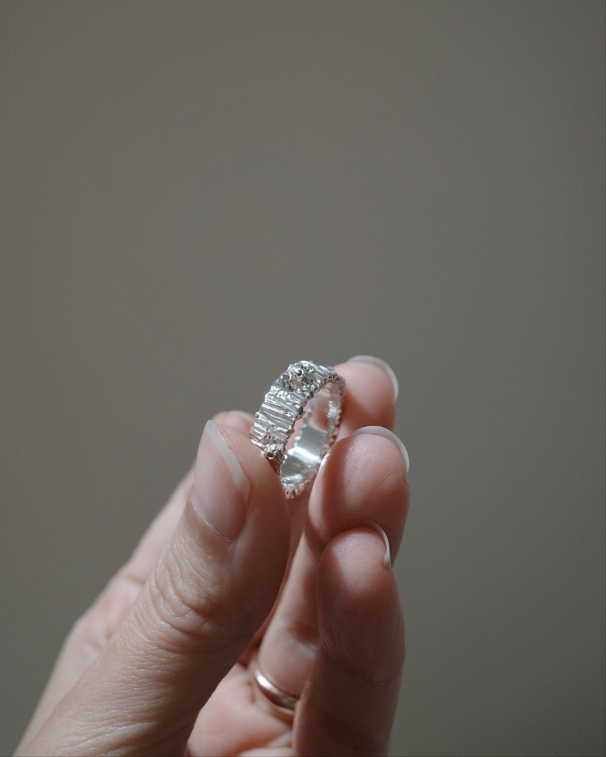 A close-up of a hand holding a silver ring with a textured, beaded outer band and a smooth inner band, set against a solid, warm gray background.